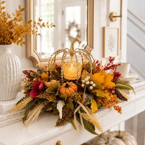 May include: A decorative gold metal pumpkin-shaped candle holder with a glass votive, surrounded by an arrangement of artificial autumn leaves, orange pumpkins, red berries, and dried wheat stalks, creating a seasonal centerpiece display.