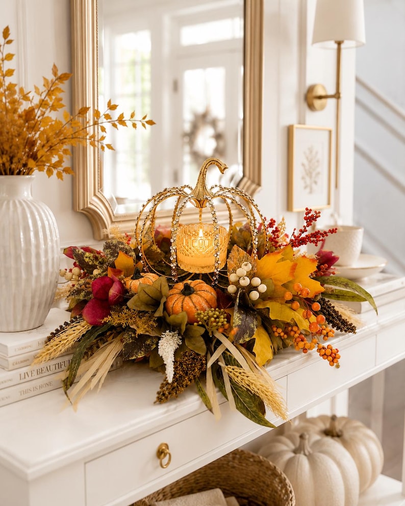 May include: A decorative gold metal pumpkin-shaped candle holder with a glass votive, surrounded by an arrangement of artificial autumn leaves, orange pumpkins, red berries, and dried wheat stalks, creating a seasonal centerpiece display.