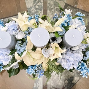 Puede incluir: Un centro de mesa con tres velas azules rodeadas de flores blancas y azules. Las flores incluyen lirios, hortensias y otras flores blancas peque&ntilde;as.