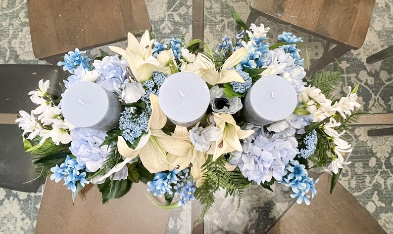 Puede incluir: Un centro de mesa con tres velas azules rodeadas de flores blancas y azules. Las flores incluyen lirios, hortensias y otras flores blancas peque&ntilde;as.