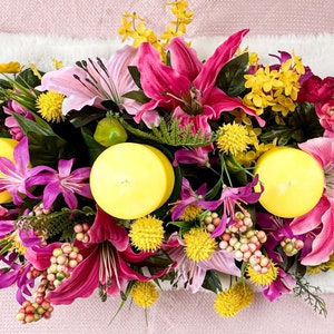 May include: A vibrant floral arrangement featuring pink lilies, yellow and purple flowers, and three yellow candles. Green leaves and small red buds add to the colorful display. The arrangement is set on a white surface.