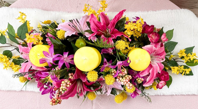 May include: A vibrant floral arrangement featuring pink lilies, yellow and purple flowers, and three yellow candles. Green leaves and small red buds add to the colorful display. The arrangement is set on a white surface.