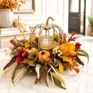 May include: A decorative centerpiece featuring a gold metal pumpkin-shaped candle holder with crystal accents, surrounded by artificial autumn foliage, orange pumpkins, red berries, wheat stalks, and cream-colored berries on a white surface.