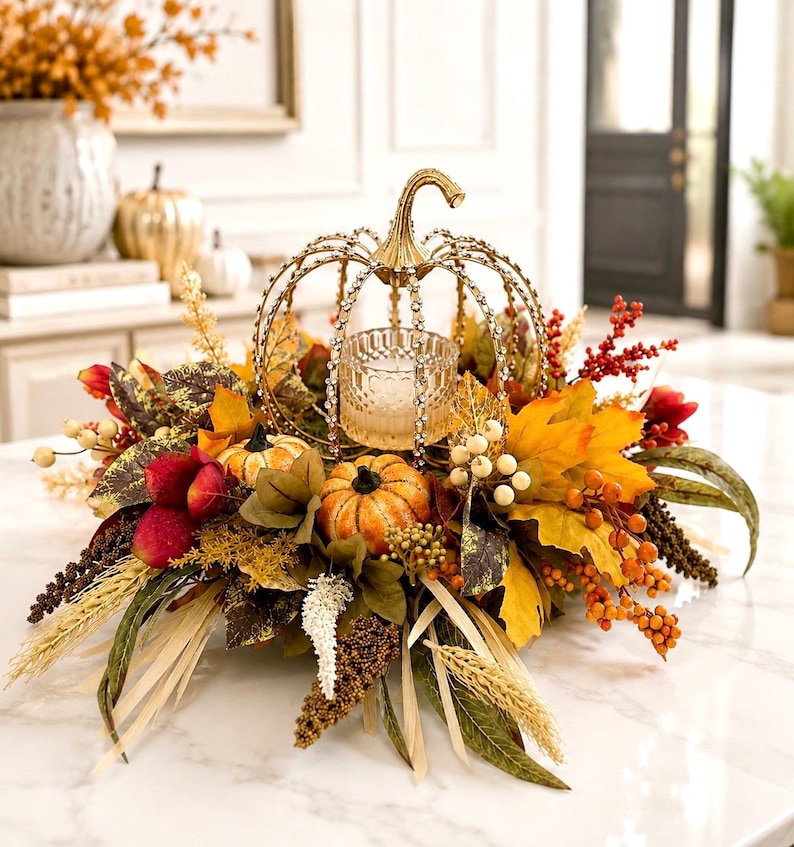 May include: A decorative centerpiece featuring a gold metal pumpkin-shaped candle holder with crystal accents, surrounded by artificial autumn foliage, orange pumpkins, red berries, wheat stalks, and cream-colored berries on a white surface.