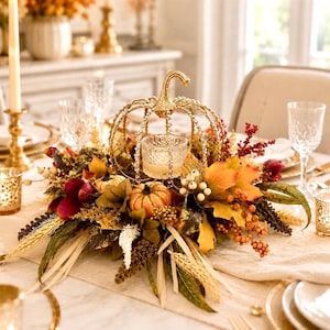 May include: A decorative gold metal pumpkin-shaped candle holder featuring a central glass votive cup, surrounded by an arrangement of artificial autumn leaves, small orange pumpkins, dried wheat stalks, and clusters of red and white berries.
