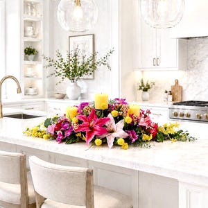 May include: A kitchen island is adorned with a vibrant floral arrangement featuring pink lilies, yellow and purple flowers, and three yellow candles. The kitchen has white cabinetry, a marble countertop, and a stainless steel stove.