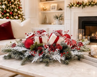 Puede incluir: Un centro de mesa navideño con ramas de pino escarchadas, adornos rojos y blancos, y dos pequeñas cajas de regalo blancas atadas con cintas rojas. El arreglo está decorado con bayas rojas y pequeños adornos en forma de caramelo.