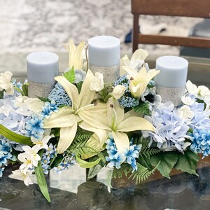 Puede incluir: Un centro de mesa con tres velas azules rodeadas de flores blancas y azules. Las velas est&aacute;n decoradas con purpurina plateada.