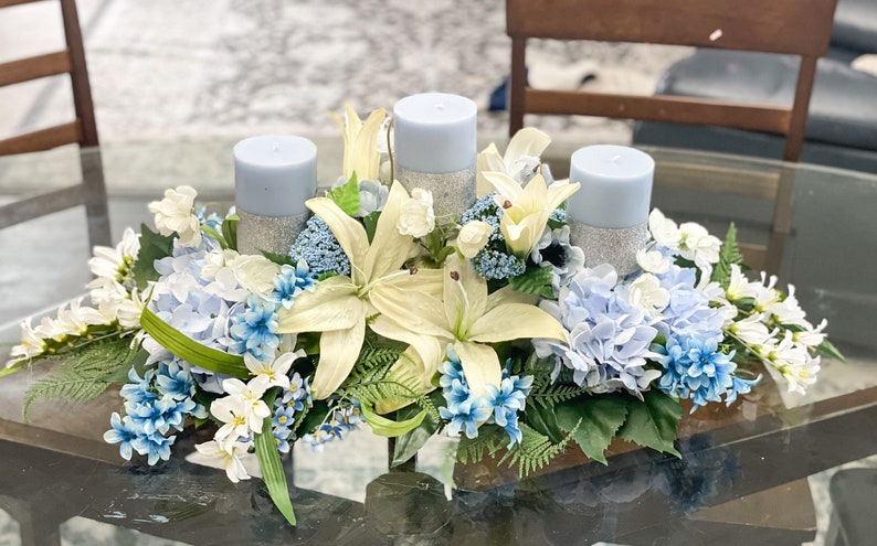 Puede incluir: Un centro de mesa con tres velas azules rodeadas de flores blancas y azules. Las velas est&aacute;n decoradas con purpurina plateada.