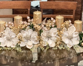 May include: A decorative centerpiece featuring four gold pillar candles surrounded by white and gold flowers, green foliage, and gold berries. The arrangement sits on a glass table, creating a festive display.