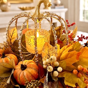 May include: A gold-toned metal pumpkin-shaped candle holder adorned with clear rhinestones, containing a faceted glass votive with a lit candle. The centerpiece is surrounded by artificial orange pumpkins, yellow and brown autumn leaves, and berries.