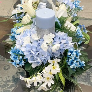 Puede incluir: Un centro de mesa floral azul y blanco con dos velas azules. Las velas tienen una banda de purpurina plateada alrededor del medio. El centro de mesa est&aacute; hecho de hortensias, lirios y otras flores.