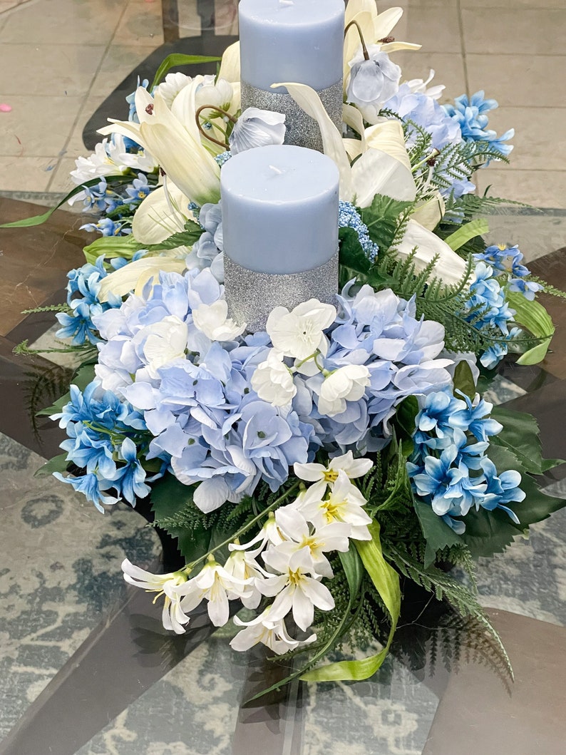Puede incluir: Un centro de mesa floral azul y blanco con dos velas azules. Las velas tienen una banda de purpurina plateada alrededor del medio. El centro de mesa est&aacute; hecho de hortensias, lirios y otras flores.