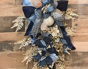 Puede incluir: Una guirnalda decorativa navideña con flores de Pascua azul marino, follaje dorado en forma de helecho y cintas en azul marino, dorado y estampados de cuadros. La guirnalda incluye adornos dorados y plateados brillantes, creando una exhibición festiva y elegante. El arreglo está sobre un fondo de madera.