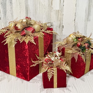 May include: Three red velvet gift boxes with gold ribbon and gold glitter accents. The boxes are decorated with gold fern sprigs and gold and red ornaments.
