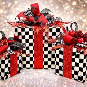Könnte beinhalten: Drei dekorative Geschenkboxen mit einem schwarz-weißen Schachbrettmuster. Jede Box ist mit einem roten Glitzerband verziert und mit roten und schwarzen Schleifen versehen. Die Boxen sind mit roten Beeren und Zweigen dekoriert.