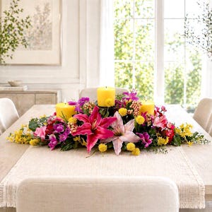 May include: A dining table centerpiece featuring a vibrant floral arrangement with pink lilies, yellow and purple flowers, and three yellow candles. The arrangement sits on a cream-colored table runner, creating an elegant display.