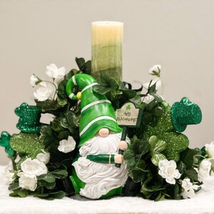 May include: A green and white gnome figurine holding a sign that says "Happy St. Patrick's Day!" The gnome is surrounded by green shamrock leaves and white flowers. A green candle is in the background.