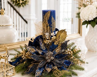 Puede incluir: Centro de mesa navideño festivo con una vela de pilar azul marino adornada con detalles de hojas doradas. El arreglo incluye una gran flor de pascua azul, elementos decorativos dorados y ramitas de hoja perenne.