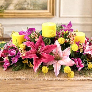 May include: A floral centerpiece with three yellow candles surrounded by pink lilies, purple orchids, and yellow flowers. The arrangement sits on a burlap runner atop a wooden table, creating a vibrant display.