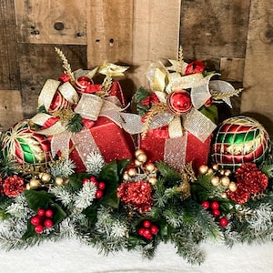 May include: A Christmas centerpiece featuring two red gift boxes with gold ribbon and green and gold ornaments, surrounded by green pine branches, red berries, and gold accents.
