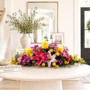 May include: A round, white table displays a vibrant floral arrangement with pink lilies, yellow candles, and various colorful flowers. A vase with greenery and books are also on the table. The setting is a bright, well-lit interior.