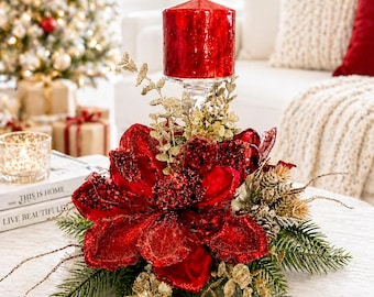Puede incluir: Centro de mesa navideño con una gran vela roja sobre un soporte de cristal, rodeada de una flor roja, detalles dorados y ramas de hoja perenne. El arreglo está sobre una mesa blanca, con un árbol de Navidad al fondo.