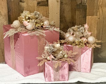 May include: Three pink velvet gift boxes with gold ribbon and embellishments. The boxes are stacked on top of each other, with the largest box on the bottom and the smallest box on top.