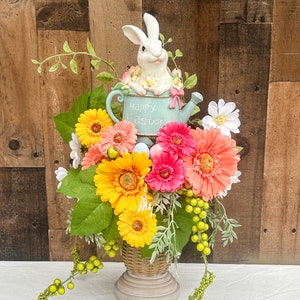 Może przedstawiać: Pastelowy niebieski konewka z białym królikiem siedzącym w środku. Na konewce widnieje napis "Happy Easter". Konewka jest otoczona różnorodnymi kolorowymi kwiatami, w tym różowymi, żółtymi i białymi gerberami. Aranżacja znajduje się w brązowym wiklinowym wazonie z jasnobrązową podstawą.