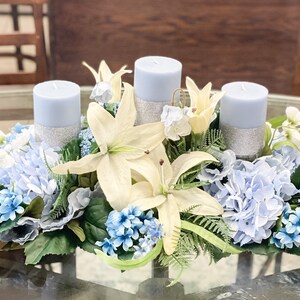 Puede incluir: Un centro de mesa con tres velas azules rodeadas de lirios blancos, hortensias azules y otras flores blancas y azules.