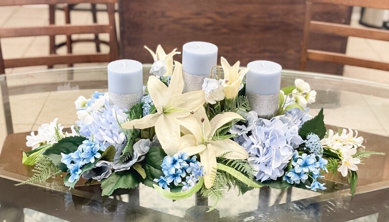 Puede incluir: Un centro de mesa con tres velas azules rodeadas de lirios blancos, hortensias azules y otras flores blancas y azules.