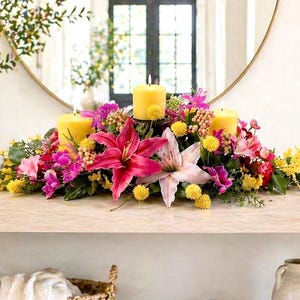 May include: A floral centerpiece with three yellow candles, surrounded by vibrant pink lilies, yellow and pink flowers, and greenery. The arrangement sits on a light-colored surface, with a mirror in the background.