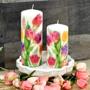 Peut inclure: Deux bougies piliers blanches décorées de motifs de tulipes à l'aquarelle rouges, roses, violettes et jaunes. Les bougies sont posées sur un plateau décoratif blanc, entourées de tulipes roses.