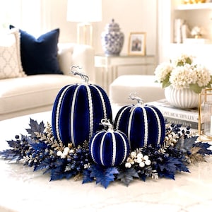 May include: Three velvet pumpkins in deep navy blue, each featuring vertical silver beaded trim and a metallic silver stem. The pumpkins are arranged on a decorative base of dark blue faux leaves, dark blue berries, and small white berries.