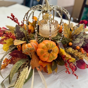 May include: A decorative centerpiece featuring a gold-toned metal pumpkin frame adorned with clear rhinestones, surrounding a glass candle holder. The arrangement includes artificial orange pumpkins, red berries, and various autumn-colored leaves.