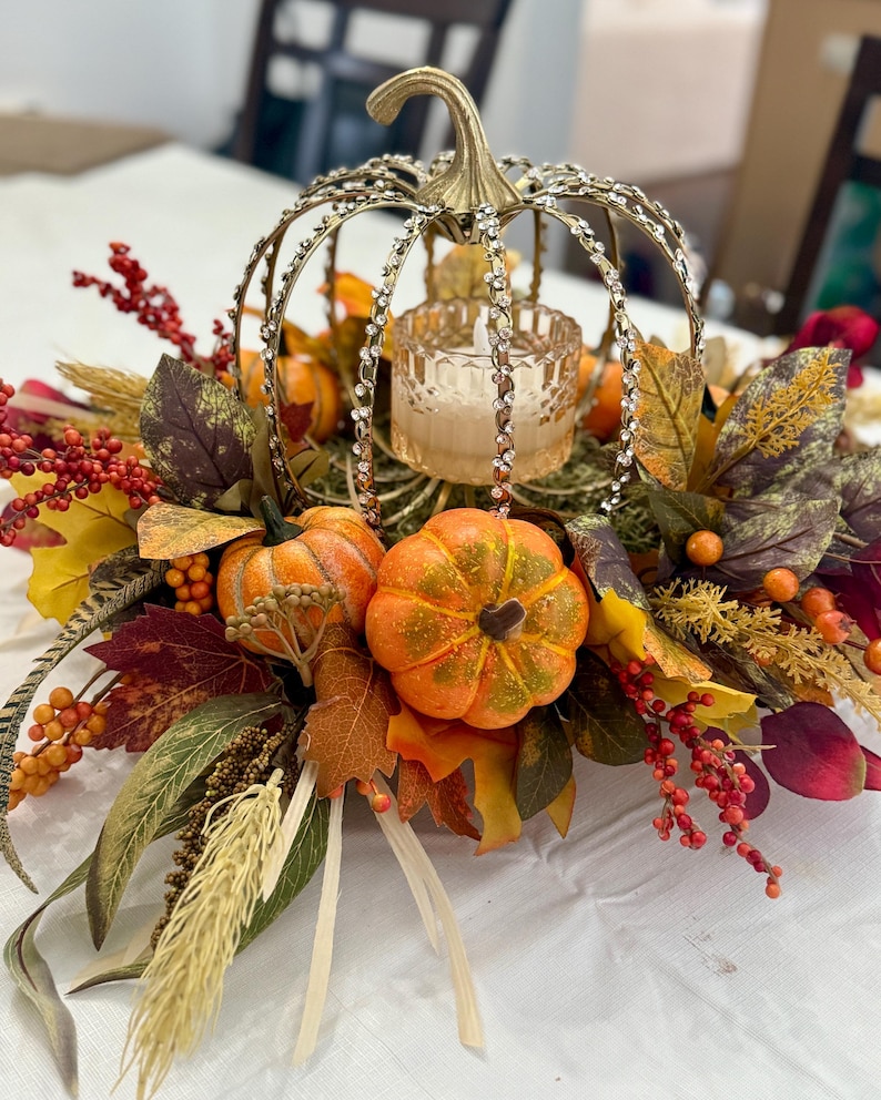 May include: A decorative centerpiece featuring a gold-toned metal pumpkin frame adorned with clear rhinestones, surrounding a glass candle holder. The arrangement includes artificial orange pumpkins, red berries, and various autumn-colored leaves.