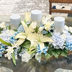 Puede incluir: Un centro de mesa con tres velas azules rodeadas de lirios blancos, hortensias azules y flores blancas. El centro de mesa est&aacute; sobre una mesa de vidrio.
