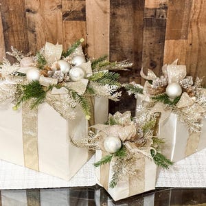 May include: Three white gift boxes with gold ribbon and embellishments, decorated with greenery and white ornaments. The boxes are arranged in a pyramid shape on a white table runner.