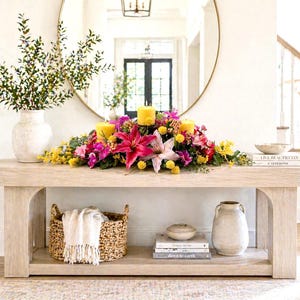 May include: A light-colored wooden console table with a floral arrangement and candles. The arrangement includes pink lilies, yellow and purple flowers, and three yellow candles. A woven basket with a white throw, books, and a vase are on the shelves.