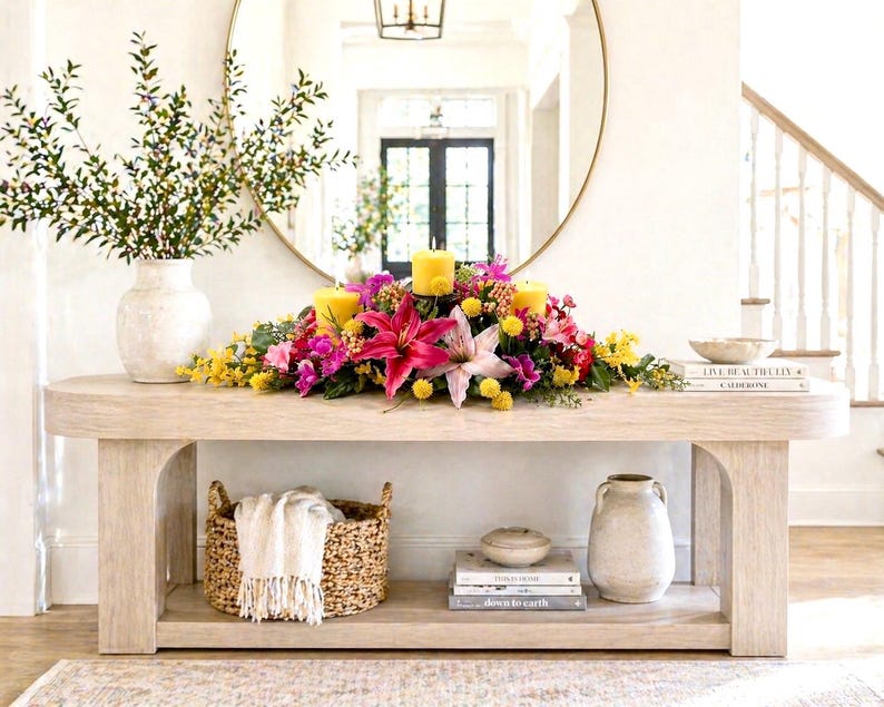 May include: A light-colored wooden console table with a floral arrangement and candles. The arrangement includes pink lilies, yellow and purple flowers, and three yellow candles. A woven basket with a white throw, books, and a vase are on the shelves.