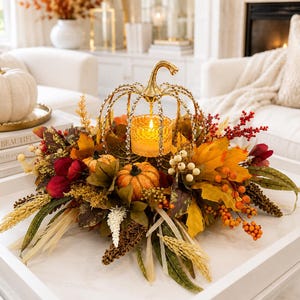 Può includere: Un centrotavola decorativo caratterizzato da una struttura a forma di zucca in metallo dorato con un portacandele centrale in vetro contenente una candela accesa, circondato da fogliame autunnale artificiale, zucche arancioni, bacche rosse e spighe di grano secche.