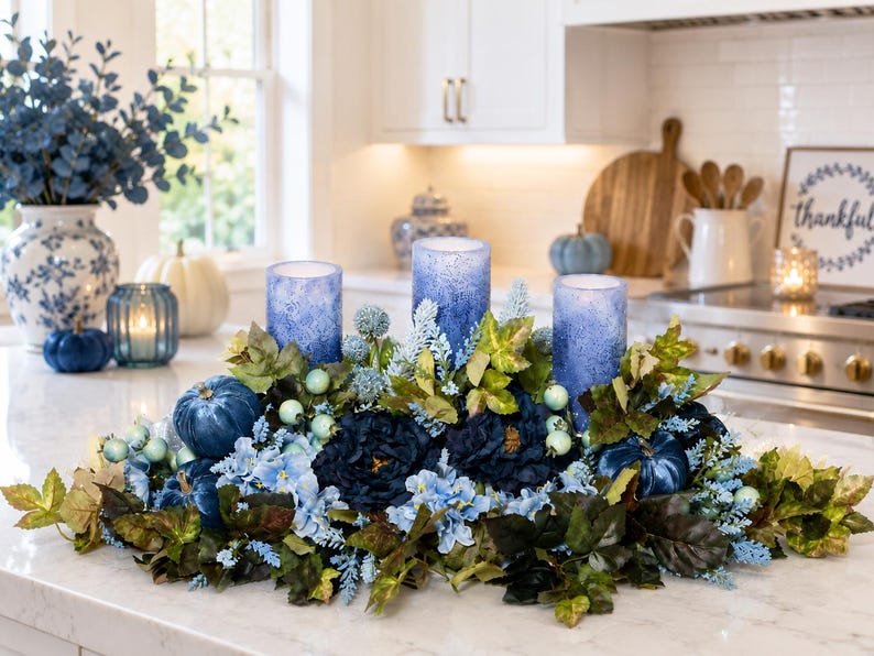 May include: A floral centerpiece featuring three blue textured pillar candles surrounded by dark blue and light blue artificial flowers, green leaves, and small blue pumpkins. The arrangement is set on a white countertop in a kitchen setting.