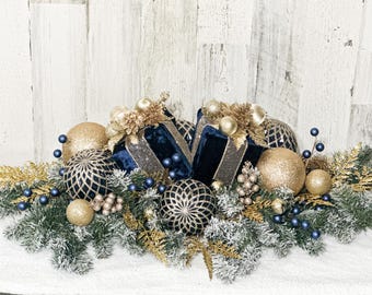 Puede incluir: Un centro de mesa navideño festivo con adornos dorados y azul marino, ramitas decorativas y una cinta azul marino. El arreglo incluye esferas brillantes y vegetación helada, creando una exhibición navideña.