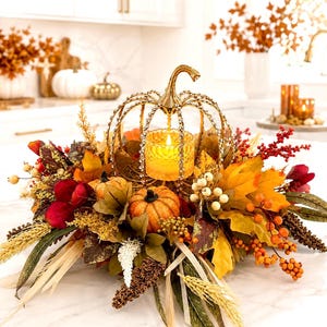 May include: A decorative centerpiece featuring a gold-toned metal pumpkin frame encrusted with clear crystals, surrounding a glowing glass candle holder. The arrangement includes artificial autumn leaves, orange pumpkins, red berries, and wheat stalks.