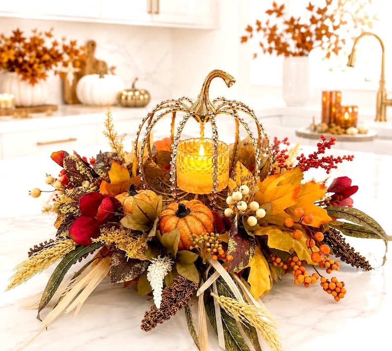 May include: A decorative centerpiece featuring a gold-toned metal pumpkin frame encrusted with clear crystals, surrounding a glowing glass candle holder. The arrangement includes artificial autumn leaves, orange pumpkins, red berries, and wheat stalks.