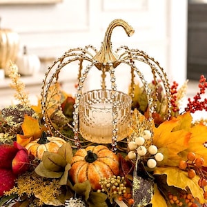 May include: A gold metal pumpkin-shaped candle holder with rhinestone accents on the ribs, containing a clear glass votive. It is surrounded by artificial autumn foliage, including orange pumpkins, yellow maple leaves, and red and white berries.