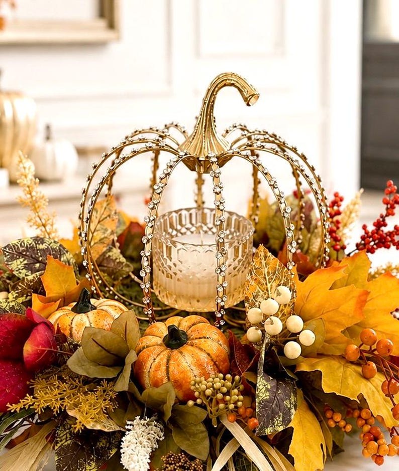 May include: A gold metal pumpkin-shaped candle holder with rhinestone accents on the ribs, containing a clear glass votive. It is surrounded by artificial autumn foliage, including orange pumpkins, yellow maple leaves, and red and white berries.
