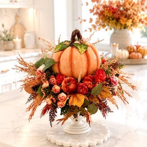 May include: A decorative centrepiece featuring a large, glossy orange ceramic pumpkin with a dark brown stem, surrounded by an arrangement of artificial red and orange roses, autumn leaves, and textured foliage, all set on a white pedestal base.
