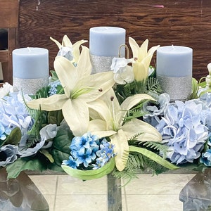 Puede incluir: Un centro de mesa con tres velas azules, lirios blancos e hortensias azules. El centro de mesa est&aacute; sobre una mesa de vidrio.