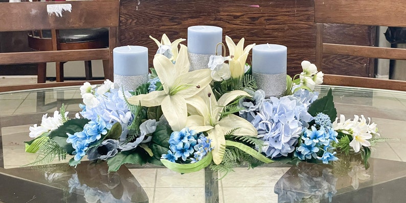 Puede incluir: Un centro de mesa con tres velas azules, lirios blancos e hortensias azules. El centro de mesa est&aacute; sobre una mesa de vidrio.